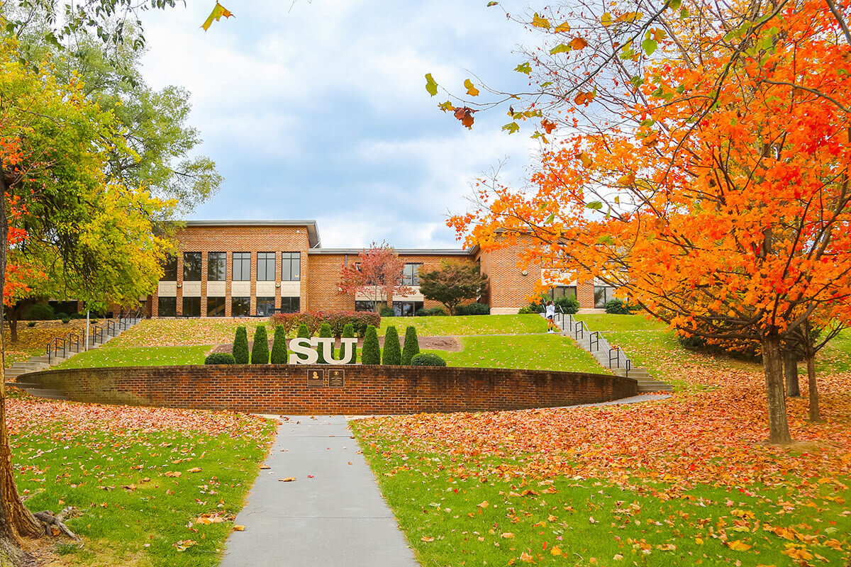  Shenandoah University in autumn, with a large brick building, trees with orange autumn leaves, and a stone wall with the letters SU in the foreground.